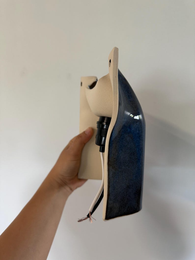 May include: A wall-mounted ceramic light fixture featuring a matte cream-colored back plate and a glossy, deep blue curved front section. The fixture includes a black socket component and exposed white and copper electrical wiring at the base.