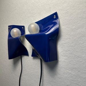 May include: Two wall-mounted lamps featuring a glossy, deep blue, folded sheet-like structure. Each lamp holds a single, spherical, frosted white light bulb. A thin black power cord extends downward from the base of each individual light fixture.
