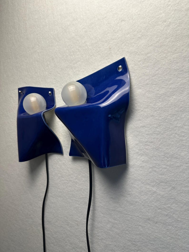 May include: Two wall-mounted lamps featuring a glossy, deep blue, folded sheet-like structure. Each lamp holds a single, spherical, frosted white light bulb. A thin black power cord extends downward from the base of each individual light fixture.