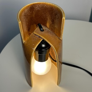 Puede incluir: Una l&aacute;mpara de mesa cil&iacute;ndrica de cer&aacute;mica con un esmalte brillante de color &aacute;mbar marr&oacute;n moteado. La estructura presenta un dise&ntilde;o curvo y superpuesto con una abertura central que alberga un casquillo negro y una bombilla esf&eacute;rica blanca esmerilada.