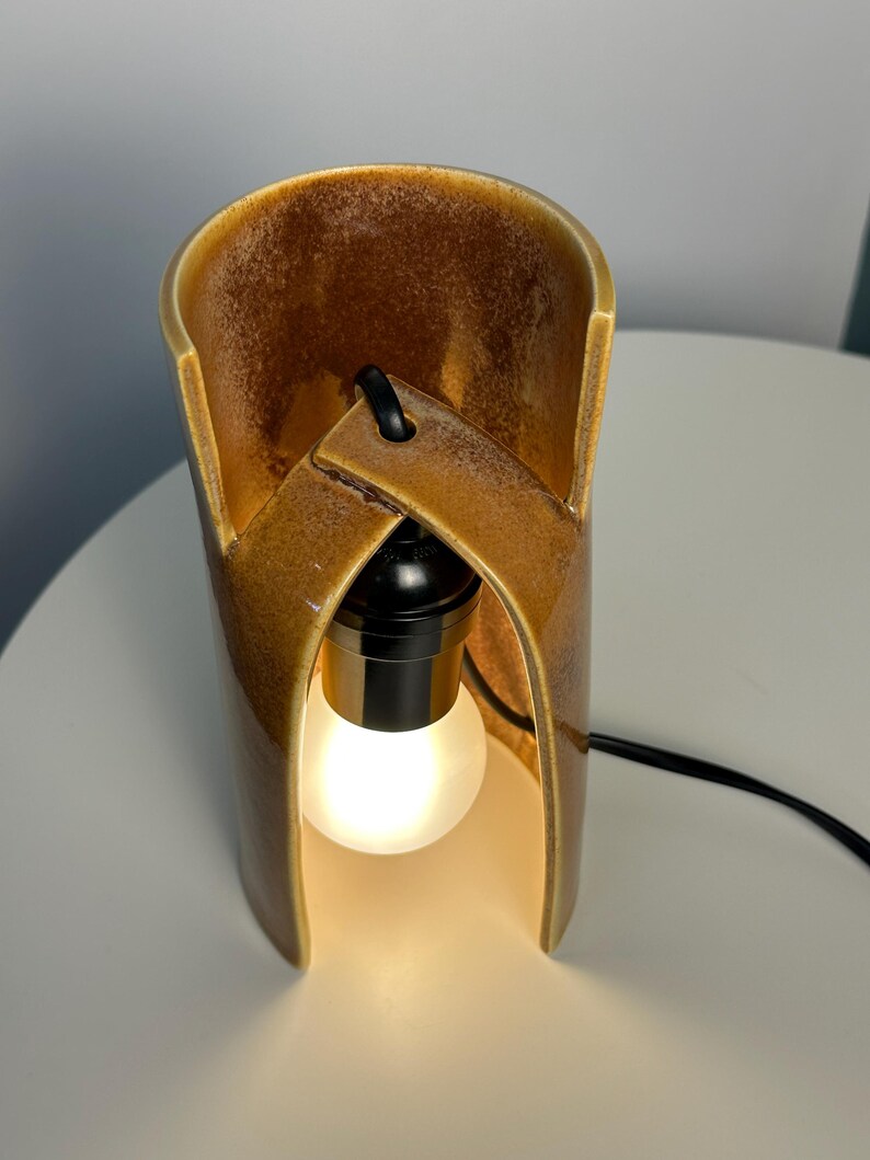 Puede incluir: Una l&aacute;mpara de mesa cil&iacute;ndrica de cer&aacute;mica con un esmalte brillante de color &aacute;mbar marr&oacute;n moteado. La estructura presenta un dise&ntilde;o curvo y superpuesto con una abertura central que alberga un casquillo negro y una bombilla esf&eacute;rica blanca esmerilada.