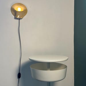 May include: A wall-mounted beige lamp with a curved, shell-like shade emitting a warm glow, featuring a black cord and switch. Below it stands a white circular side table with a flat top surface and a deep, cylindrical storage compartment.