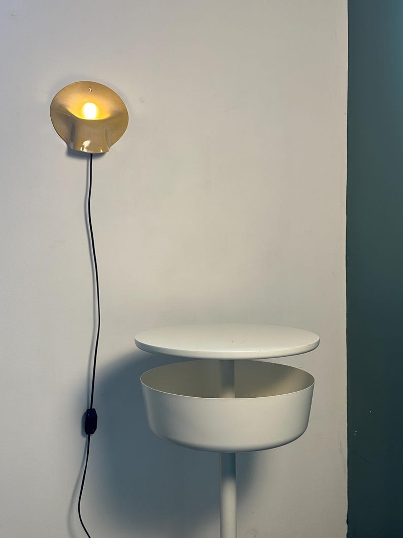 May include: A wall-mounted beige lamp with a curved, shell-like shade emitting a warm glow, featuring a black cord and switch. Below it stands a white circular side table with a flat top surface and a deep, cylindrical storage compartment.