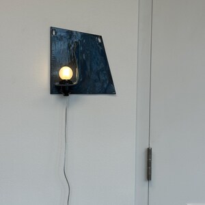 May include: A modern wall sconce with a dark blue, irregularly shaped metal shade. A round, illuminated light bulb is mounted on a black fixture. A silver cord extends downward. The sconce is mounted on a white wall.