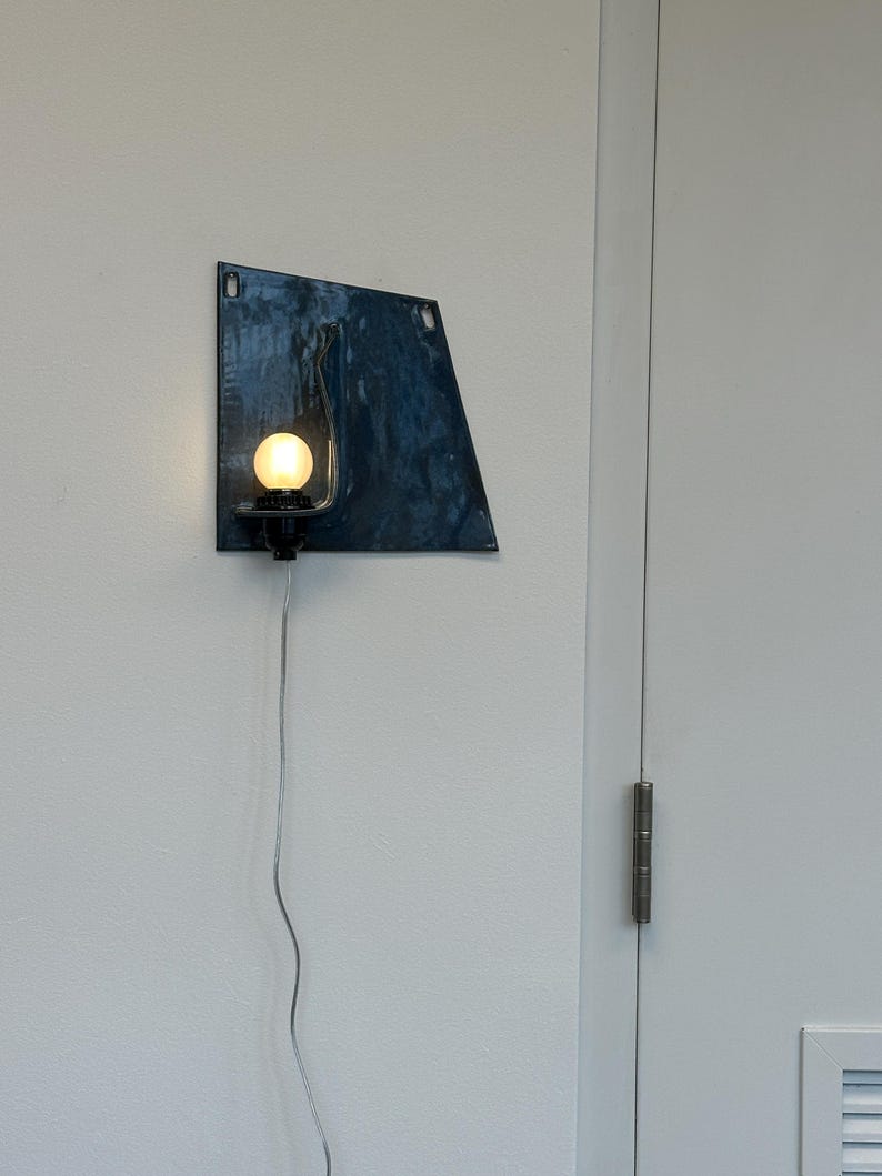 May include: A modern wall sconce with a dark blue, irregularly shaped metal shade. A round, illuminated light bulb is mounted on a black fixture. A silver cord extends downward. The sconce is mounted on a white wall.