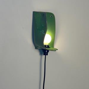 Pu&ograve; includere: Una lampada da parete con un paralume in ceramica verde lucido e curvo dalla finitura liscia. Una singola lampadina bianca rotonda e luminosa &egrave; posizionata al centro del paralume, collegata a un portalampada nero e a un lungo cavo elettrico nero.