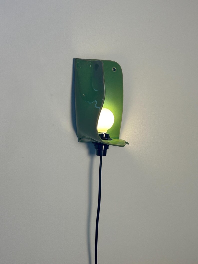 Pu&ograve; includere: Una lampada da parete con un paralume in ceramica verde lucido e curvo dalla finitura liscia. Una singola lampadina bianca rotonda e luminosa &egrave; posizionata al centro del paralume, collegata a un portalampada nero e a un lungo cavo elettrico nero.