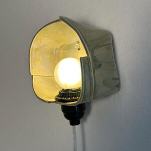 Puede incluir: Un aplique de pared con una pantalla de cer&aacute;mica curva de color verde p&aacute;lido y acabado brillante. Una bombilla blanca redonda y brillante se sit&uacute;a en el centro de la pantalla, unida a un casquillo negro con un cable transparente hacia abajo.