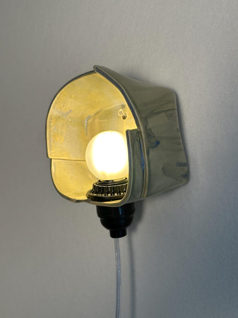 Puede incluir: Un aplique de pared con una pantalla de cer&aacute;mica curva de color verde p&aacute;lido y acabado brillante. Una bombilla blanca redonda y brillante se sit&uacute;a en el centro de la pantalla, unida a un casquillo negro con un cable transparente hacia abajo.