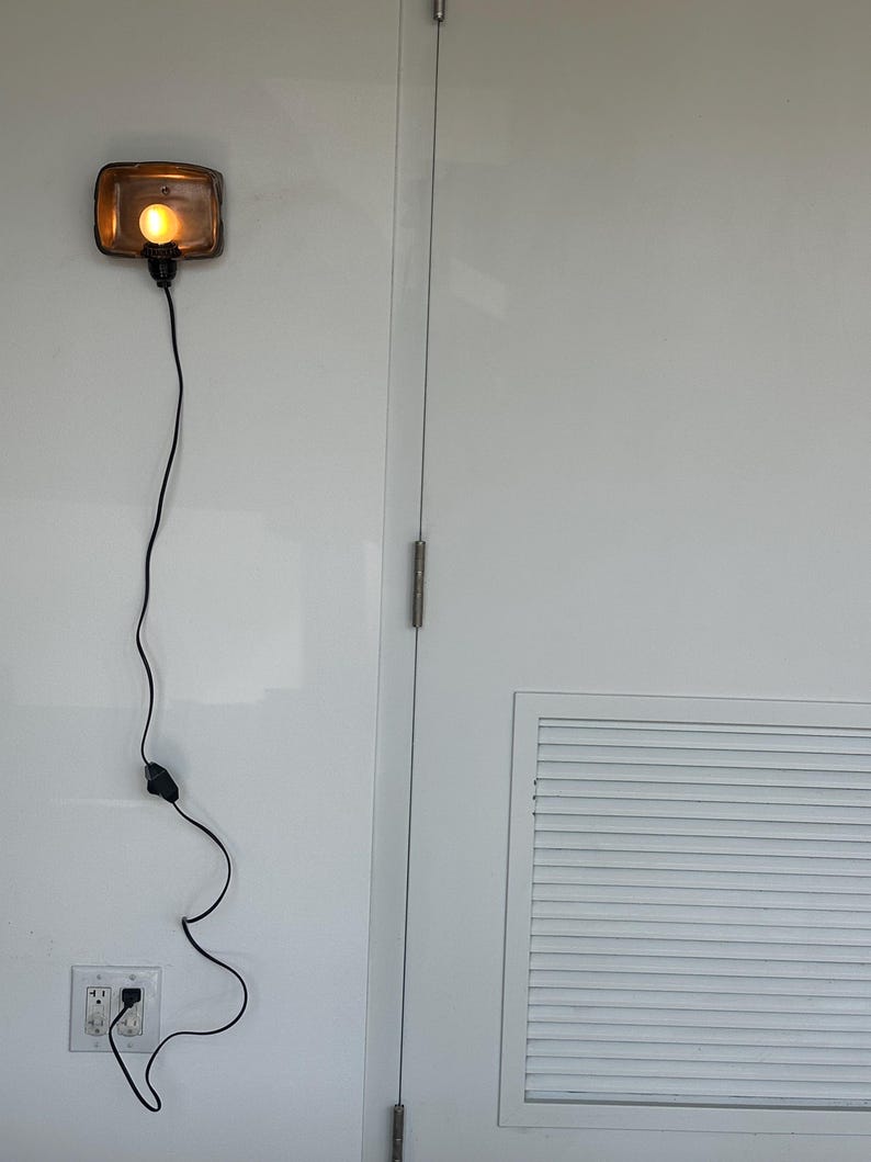 May include: A rectangular, amber-colored wall-mounted light fixture with a visible bulb, connected by a black cord to a wall outlet. The light is mounted on a white wall next to a white door.