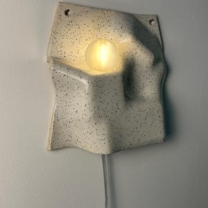May include: A wall-mounted ceramic light fixture with an off-white, speckled finish and an irregular, wavy surface. A single, round, frosted light bulb is recessed into the center of the form, emitting a warm glow. A clear power cord hangs below.