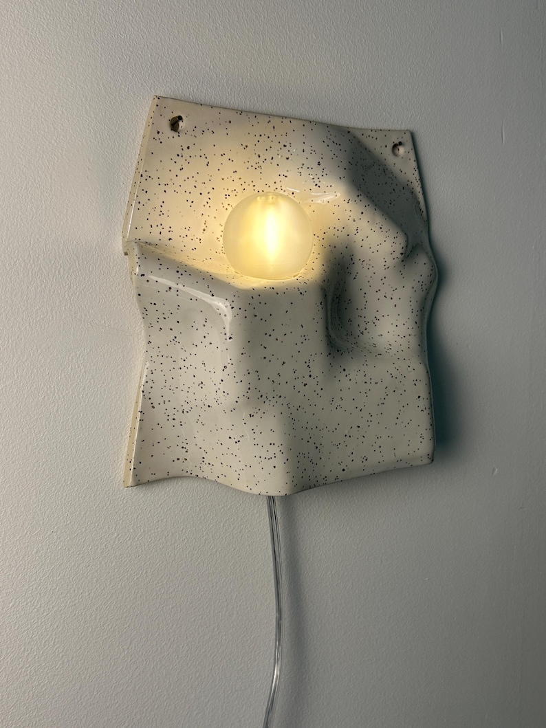 May include: A wall-mounted ceramic light fixture with an off-white, speckled finish and an irregular, wavy surface. A single, round, frosted light bulb is recessed into the center of the form, emitting a warm glow. A clear power cord hangs below.