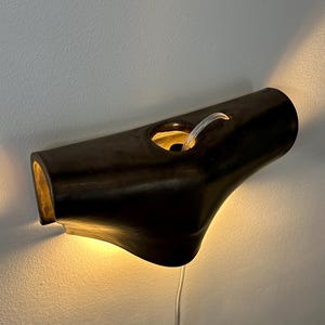 May include: A dark brown, horizontal wall-mounted light fitting with a smooth, curved, tubular shape. The fitting features a central circular opening with a visible internal light source that casts a warm glow against the wall below and behind it.