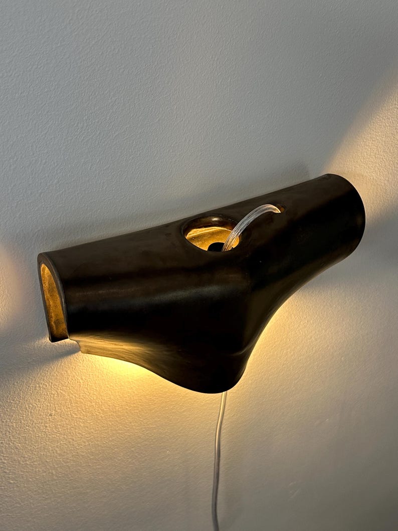May include: A dark brown, horizontal wall-mounted light fitting with a smooth, curved, tubular shape. The fitting features a central circular opening with a visible internal light source that casts a warm glow against the wall below and behind it.