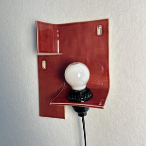 Pode incluir: Arandela de parede moderna com abajur de cer&acirc;mica vermelha e l&acirc;mpada branca. A base e o cabo s&atilde;o pretos. O design &eacute; minimalista e contempor&acirc;neo.