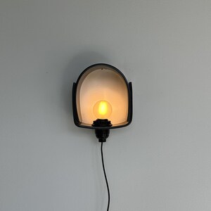 Puede incluir: Una l&aacute;mpara de pared negra con una bombilla c&aacute;lida y brillante. La l&aacute;mpara tiene un dise&ntilde;o curvo y arqueado con un marco negro y un interior de color claro. Un cable negro se extiende hacia abajo.