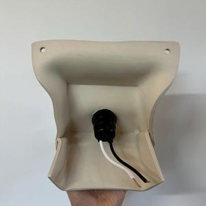 May include: A beige wall-mounted light fixture base with a curved, flared shape. It features two circular mounting holes at the top corners and a central black socket component with one white and one black electrical wire extending from the center.