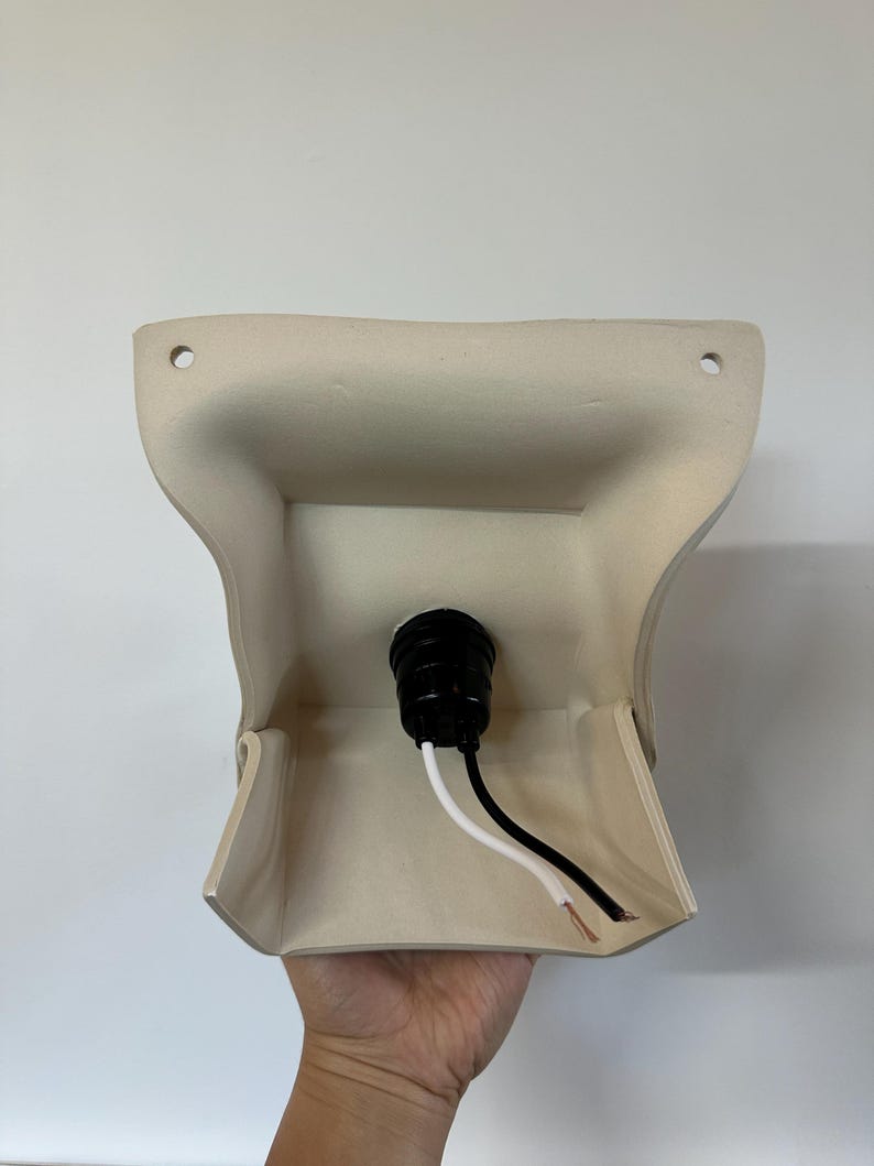 May include: A beige wall-mounted light fixture base with a curved, flared shape. It features two circular mounting holes at the top corners and a central black socket component with one white and one black electrical wire extending from the center.