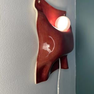 K&ouml;nnte beinhalten: Eine Wandleuchte aus Keramik mit einer gl&auml;nzenden, tiefroten Glasur und einer organisch gefalteten Form. Die Leuchte enth&auml;lt eine leuchtende, wei&szlig;e, kugelf&ouml;rmige Gl&uuml;hbirne und ein d&uuml;nnes, transparentes Stromkabel an der Unterseite.