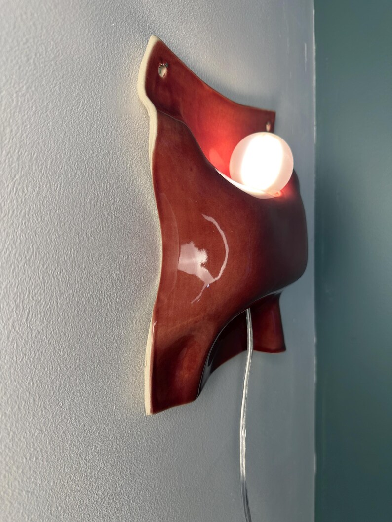 K&ouml;nnte beinhalten: Eine Wandleuchte aus Keramik mit einer gl&auml;nzenden, tiefroten Glasur und einer organisch gefalteten Form. Die Leuchte enth&auml;lt eine leuchtende, wei&szlig;e, kugelf&ouml;rmige Gl&uuml;hbirne und ein d&uuml;nnes, transparentes Stromkabel an der Unterseite.