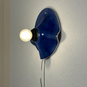 May include: A blue ceramic wall sconce with a wavy design. The sconce has a light bulb and a black base. A silver cord extends from the bottom of the sconce. The sconce is mounted on a gray wall.