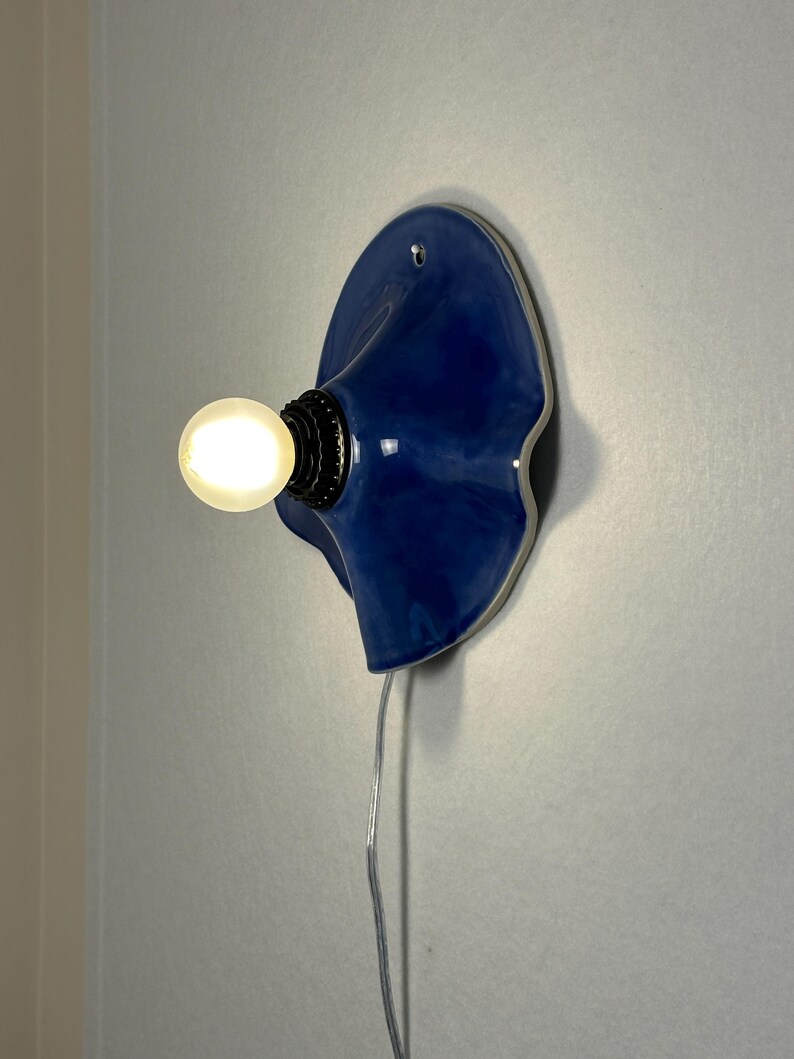 May include: A blue ceramic wall sconce with a wavy design. The sconce has a light bulb and a black base. A silver cord extends from the bottom of the sconce. The sconce is mounted on a gray wall.