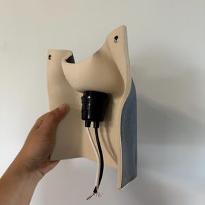 May include: A wall-mounted light fixture featuring a curved, matte beige ceramic shade with a dark blue exterior finish. The fixture includes a central black socket housing with two exposed electrical wires, one black and one white, extending downward.
