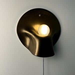 May include: A wall-mounted sconce featuring a dark, matte, organic-shaped metal shade with a curved, recessed center. A single, glowing, frosted white spherical light bulb is positioned within the indentation. A thin, clear power cord hangs below.