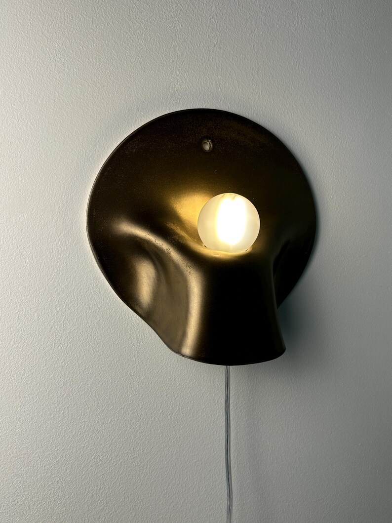 May include: A wall-mounted sconce featuring a dark, matte, organic-shaped metal shade with a curved, recessed center. A single, glowing, frosted white spherical light bulb is positioned within the indentation. A thin, clear power cord hangs below.