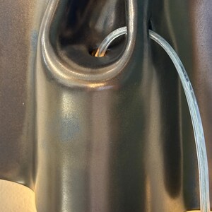 May include: A dark brown, smooth, organic-shaped ceramic wall sconce with a central opening. A clear, flexible plastic cord emerges from the center of the fixture, which emits a warm, soft light from the bottom edge against the wall surface.