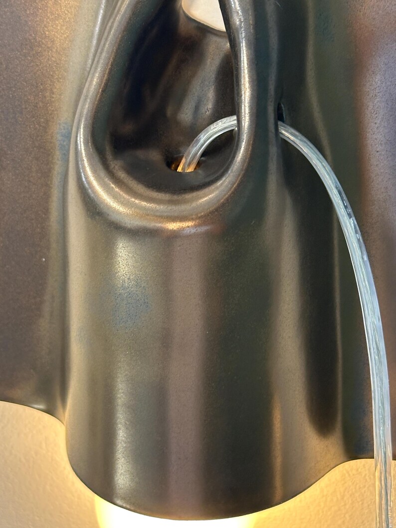 May include: A dark brown, smooth, organic-shaped ceramic wall sconce with a central opening. A clear, flexible plastic cord emerges from the center of the fixture, which emits a warm, soft light from the bottom edge against the wall surface.