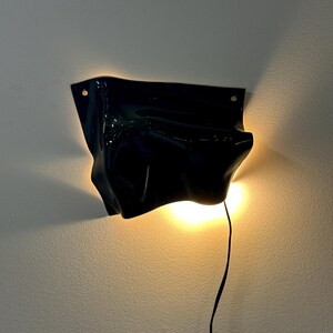 Puede incluir: Un aplique de pared negro brillante con una forma irregular, fluida y org&aacute;nica. La l&aacute;mpara emite un brillo c&aacute;lido y suave desde detr&aacute;s de sus bordes curvos, proyectando luz sobre la pared. Un fino cable de alimentaci&oacute;n negro cuelga.