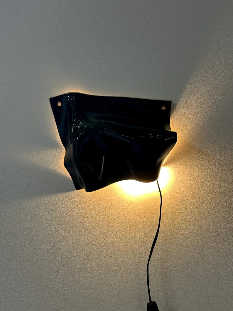 Puede incluir: Un aplique de pared negro brillante con una forma irregular, fluida y org&aacute;nica. La l&aacute;mpara emite un brillo c&aacute;lido y suave desde detr&aacute;s de sus bordes curvos, proyectando luz sobre la pared. Un fino cable de alimentaci&oacute;n negro cuelga.