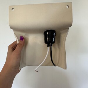 Peut inclure: Une base de luminaire murale beige pr&eacute;sentant une forme rectangulaire incurv&eacute;e avec deux trous de montage circulaires en haut. Une douille en plastique noir est centr&eacute;e sur le devant, avec un fil blanc et un fil noir d&eacute;passant vers le bas.