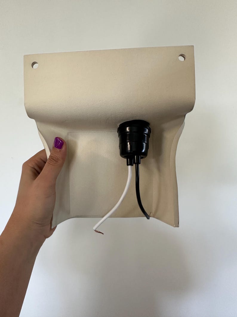 Peut inclure: Une base de luminaire murale beige pr&eacute;sentant une forme rectangulaire incurv&eacute;e avec deux trous de montage circulaires en haut. Une douille en plastique noir est centr&eacute;e sur le devant, avec un fil blanc et un fil noir d&eacute;passant vers le bas.