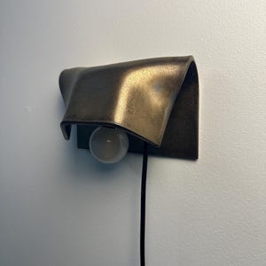Pode incluir: Um candeeiro de parede com um abajur de metal curvo em tom de bronze com acabamento texturizado. Uma l&acirc;mpada esf&eacute;rica branca est&aacute; posicionada sob o abajur e um fio de alimenta&ccedil;&atilde;o preto fino pende verticalmente da base do candeeiro.