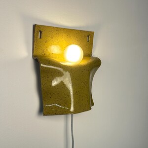 May include: A wall-mounted ceramic sconce featuring a glossy, mustard-yellow glaze with dark speckles. The fixture has a curved, folded sheet-like form with two rectangular mounting holes at the top and a single glowing spherical light bulb.