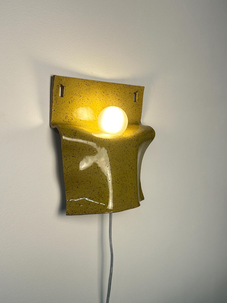 May include: A wall-mounted ceramic sconce featuring a glossy, mustard-yellow glaze with dark speckles. The fixture has a curved, folded sheet-like form with two rectangular mounting holes at the top and a single glowing spherical light bulb.