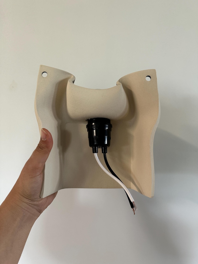 May include: A beige ceramic wall sconce fixture with a curved, molded shape featuring two circular mounting holes at the top corners. A black plastic light socket is centered in the middle, with one white and one black electrical wire extending.