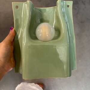 May include: A sage green ceramic wall sconce with a glossy finish and a wavy, organic shape. The fixture features a central circular opening holding a frosted white glass globe light bulb and two small holes at the top for mounting purposes.
