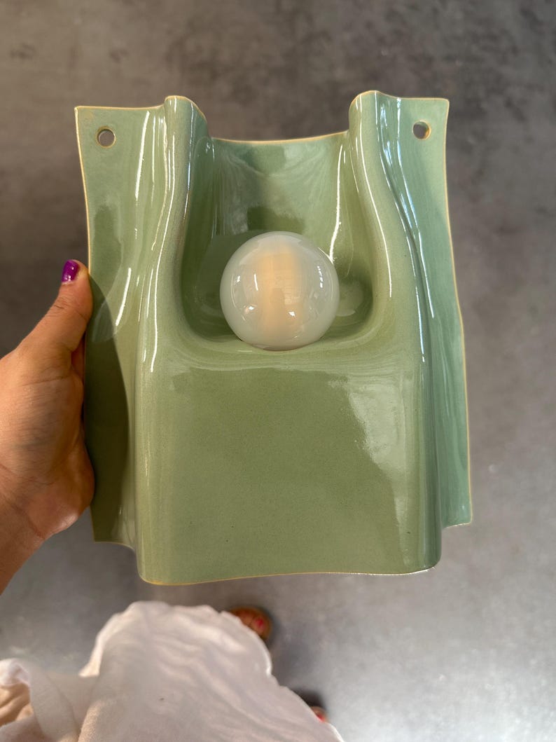 May include: A sage green ceramic wall sconce with a glossy finish and a wavy, organic shape. The fixture features a central circular opening holding a frosted white glass globe light bulb and two small holes at the top for mounting purposes.