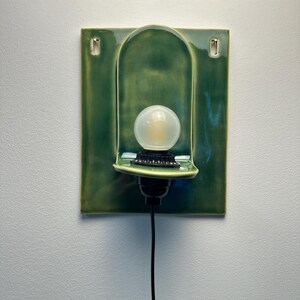 May include: A wall-mounted ceramic sconce featuring a glossy, deep green rectangular backplate with a central arched indentation. A black socket holds a round, frosted white light bulb, and a thin black power cord extends downward from the base.