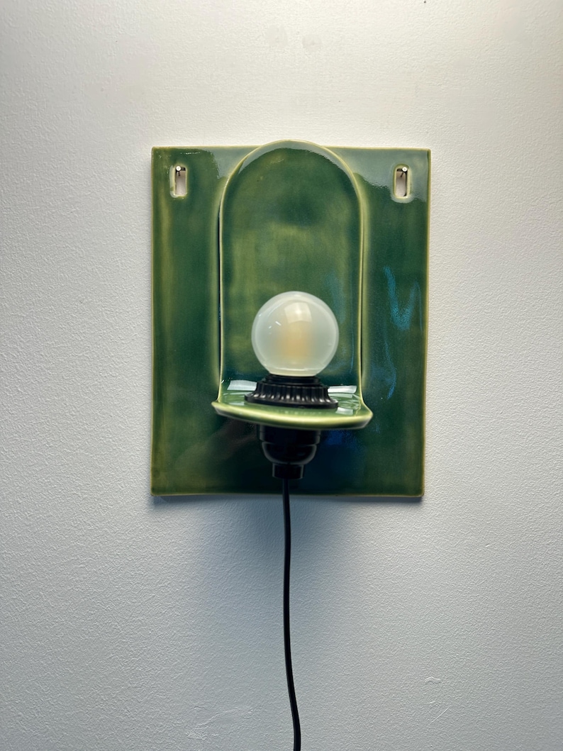 May include: A wall-mounted ceramic sconce featuring a glossy, deep green rectangular backplate with a central arched indentation. A black socket holds a round, frosted white light bulb, and a thin black power cord extends downward from the base.