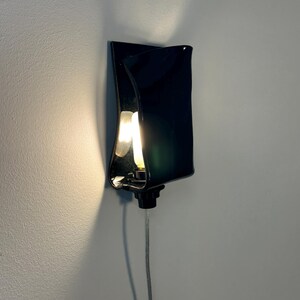 May include: A wall-mounted lamp featuring a glossy black, curved rectangular shade that partially conceals a glowing light bulb. The fixture includes a black base and a clear power cord extending downward, casting a warm light against the wall.