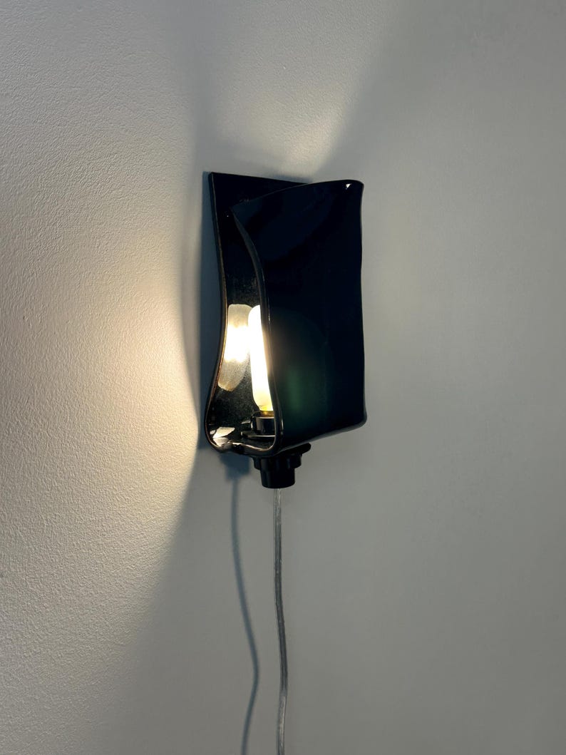 May include: A wall-mounted lamp featuring a glossy black, curved rectangular shade that partially conceals a glowing light bulb. The fixture includes a black base and a clear power cord extending downward, casting a warm light against the wall.