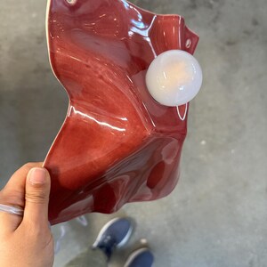 Puede incluir: Un aplique de pared con una pantalla de cer&aacute;mica de color rojo intenso brillante con una superficie irregular, ondulada y plegada. La pantalla tiene dos orificios de montaje circulares en las esquinas superiores y una bombilla esf&eacute;rica blanca.