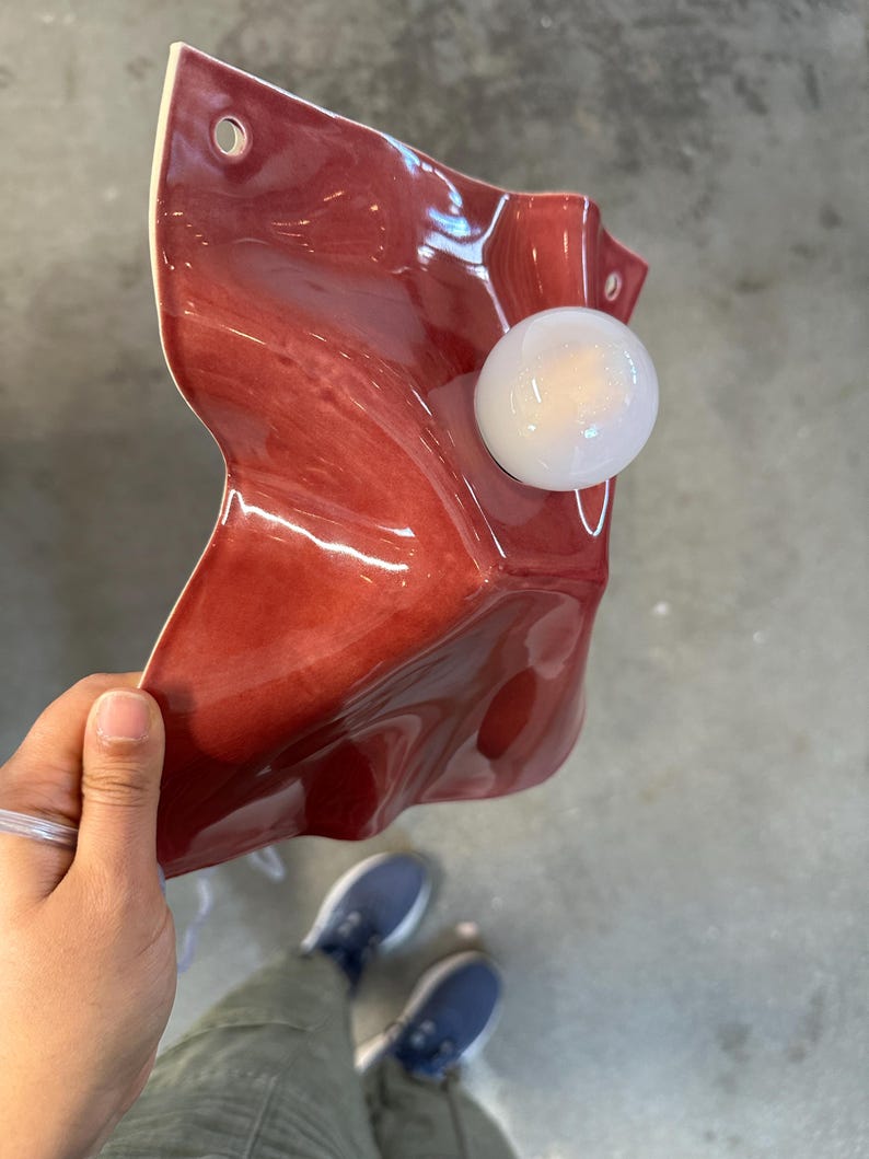 Puede incluir: Un aplique de pared con una pantalla de cer&aacute;mica de color rojo intenso brillante con una superficie irregular, ondulada y plegada. La pantalla tiene dos orificios de montaje circulares en las esquinas superiores y una bombilla esf&eacute;rica blanca.