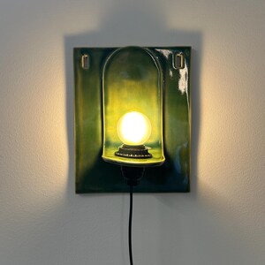 May include: A rectangular wall sconce featuring a glossy, deep green ceramic backplate with a central arched alcove. A single round, glowing light bulb is mounted within the alcove, connected to a black power cord extending downward.