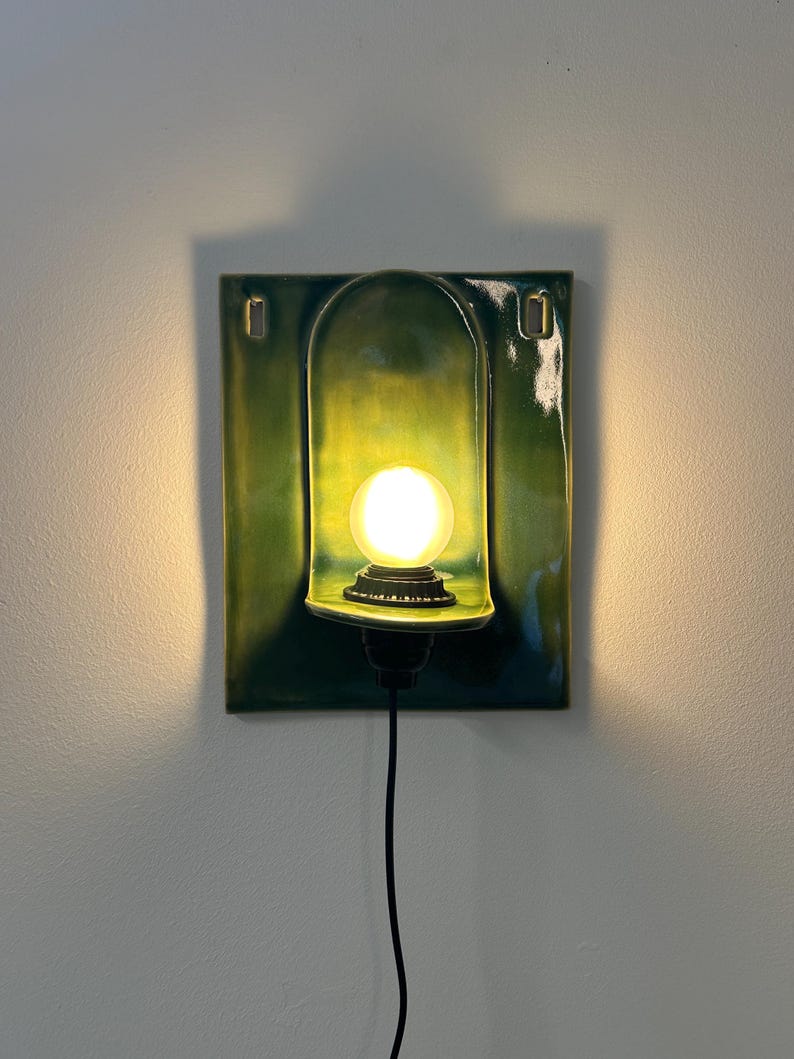 May include: A rectangular wall sconce featuring a glossy, deep green ceramic backplate with a central arched alcove. A single round, glowing light bulb is mounted within the alcove, connected to a black power cord extending downward.