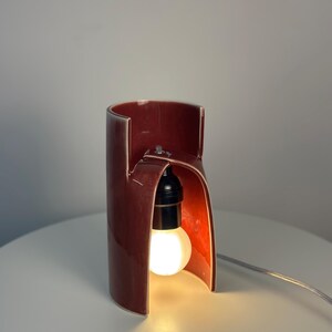 May include: A cylindrical ceramic table lamp with a glossy dark red finish. The lamp features a cutout arch design that exposes a black socket holding a glowing light bulb. A thin, light-colored electrical cord extends from the base of the lamp.